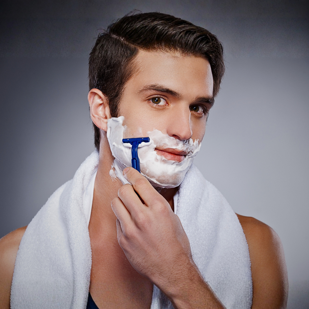 A handsome young gentleman with dark hair is shaving with a hand razor with a white towel around his neck. ©2025 Kandilynns.com